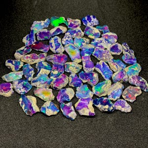 May include: A pile of raw opal gemstones, showcasing a spectrum of colours such as blue, purple, green, and red. The opals have a rough, natural texture and are spread across a dark surface.