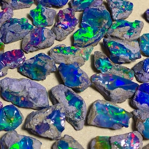 以下が含まれることがあります： 原石のオパールが多数写っています。青、緑、紫など様々な色が混ざり合い、グレーと茶色の岩石に囲まれています。石のサイズや形は様々で、自然な未研磨の風合いが特徴です。