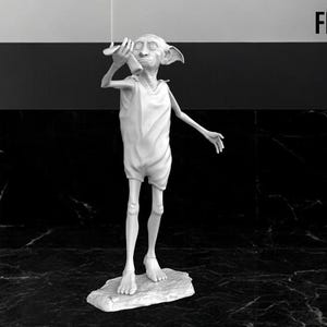 Może przedstawiać: Biała figurka Dobby'ego, domowego skrzata z serii o Harrym Potterze. Dobby stoi na małej podstawie i trzyma skarpetkę w prawej ręce. Figurka jest w neutralnej pozie z wyciągniętymi rękami.