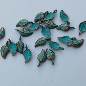 Peut inclure: Une collection de petits charmes en forme de feuilles. Chaque charme pr&eacute;sente une feuille de couleur sarcelle avec des veines et des bords de couleur bronze. Les charmes sont dispers&eacute;s sur une surface blanche, mettant en valeur leurs d&eacute;tails complexes et leur finition m&eacute;tallique.