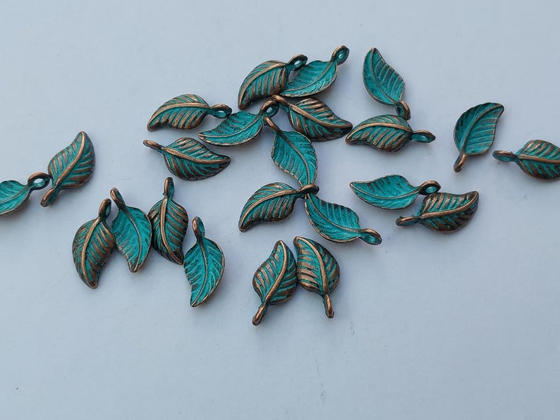 Peut inclure: Une collection de petits charmes en forme de feuilles. Chaque charme pr&eacute;sente une feuille de couleur sarcelle avec des veines et des bords de couleur bronze. Les charmes sont dispers&eacute;s sur une surface blanche, mettant en valeur leurs d&eacute;tails complexes et leur finition m&eacute;tallique.