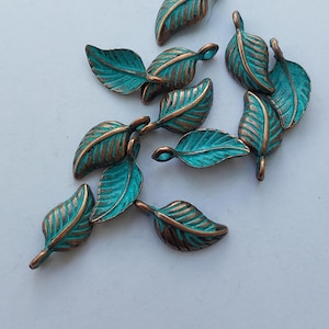 Peut inclure: Un ensemble de petits charmes d&eacute;coratifs en forme de feuilles. Chaque feuille pr&eacute;sente une surface de couleur turquoise avec des veines et des bords de couleur bronze. Les feuilles sont dispos&eacute;es sur une surface blanche, mettant en valeur leurs d&eacute;tails.