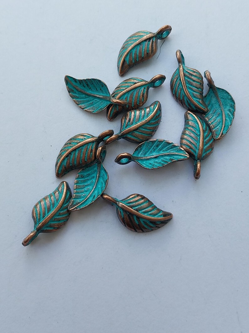 Peut inclure: Un ensemble de petits charmes d&eacute;coratifs en forme de feuilles. Chaque feuille pr&eacute;sente une surface de couleur turquoise avec des veines et des bords de couleur bronze. Les feuilles sont dispos&eacute;es sur une surface blanche, mettant en valeur leurs d&eacute;tails.