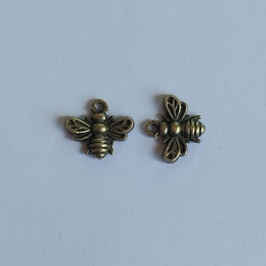 May include: Two bronze-colored bee charms. Each charm features detailed wings, body segments, and a small loop at the top for attaching to jewelry or other crafts. The bees are positioned against a light background.