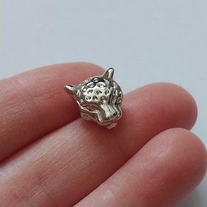 May include: A small, silver-coloured charm shaped like a jaguar's head. The charm has detailed features, including textured spots and facial features. The jaguar's head is shown in a close-up shot, held in a person's hand.