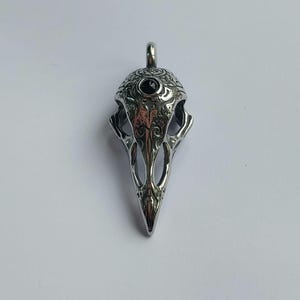 May include: Silver pendant shaped like a bird skull with intricate detailing and a black gemstone eye.