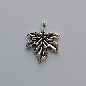 May include: A silver-toned leaf pendant with detailed veining. The pendant is shaped like a maple leaf, featuring a loop at the top for attaching to a chain. The leaf has a textured surface and is set against a white backdrop.