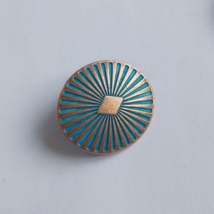Op de afbeelding: Een ronde, koperkleurige knoop met een blauw emaille ontwerp. De knoop heeft een zonnestralenpatroon met een diamant in het midden.