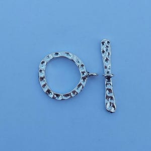May include: A silver-colored toggle clasp with a hammered texture. The clasp consists of a circular ring and a bar, both with a textured surface. The ring has a small loop attached for connection. The bar has a rounded end.