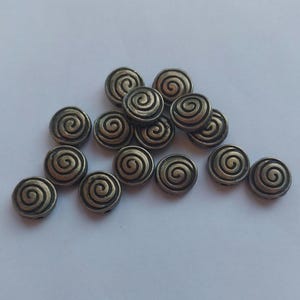 May include: A collection of small, round metal buttons featuring a raised spiral design in a dark, antique bronze finish. Each button has a circular shape with a centred, embossed swirl pattern that creates a textured, metallic appearance.