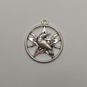May include: A silver-toned metal pendant featuring a circular frame enclosing a five-pointed star, or pentagram, with a detailed raven perched in the center. The design includes small crescent moon accents on the sides and a small jump ring at the top.