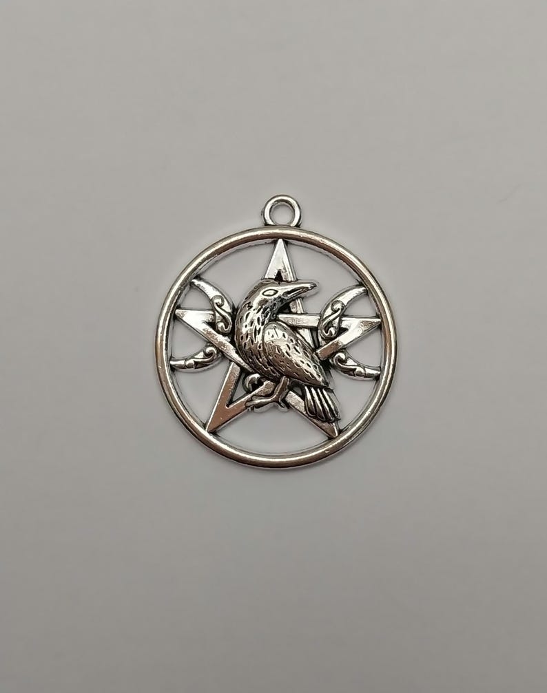 May include: A silver-toned metal pendant featuring a circular frame enclosing a five-pointed star, or pentagram, with a detailed raven perched in the center. The design includes small crescent moon accents on the sides and a small jump ring at the top.