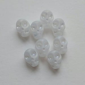 May include: A collection of eight frosted glass skull beads. Each bead has detailed facial features, including eye sockets, a nose cavity, and a jawline. The skulls are a pale, translucent white.