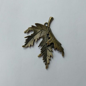 Pode incluir: Um pingente de folha de metal bronze com detalhes intrincados de veias. A folha tem uma superfície texturizada e um pequeno laço na parte superior para pendurar. O pingente é provavelmente para fazer joias ou artesanato.
