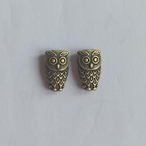 May include: Two small, identical owl-shaped charms or beads made of a metallic material with an antique bronze finish. Each owl features detailed engravings of large circular eyes, pointed ear tufts, and a textured pattern representing feathers.