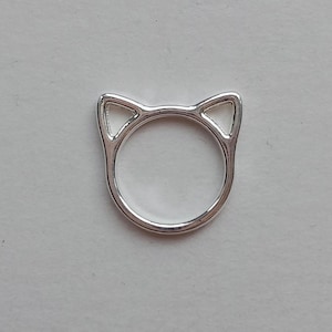 Puede incluir: Un anillo plateado con diseño de orejas de gato. El anillo tiene una banda circular con dos orejas de gato triangulares en la parte superior. El anillo está hecho de un material metálico brillante y está sobre un fondo claro. Es una joya.