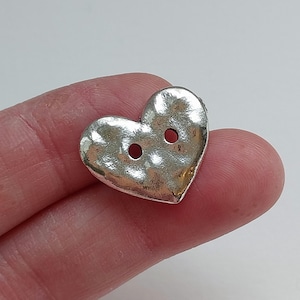 Può includere: Un bottone a forma di cuore color argento con due fori. Il bottone ha una superficie strutturata e un aspetto leggermente martellato. Ideale per progetti di cucito, artigianato o per aggiungere un tocco decorativo a vestiti o accessori.