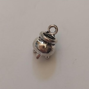 Könnte beinhalten: Ein kleiner, silberfarbener Metallanhänger in Form eines dreibeinigen Kessels. Der Anhänger hat einen abgerundeten Körper, einen breiten Rand und eine kreisförmige Öse an der Oberseite zur Befestigung. Die Oberfläche ist poliert und reflektierend.