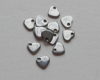 May include: A collection of small, shiny, silver-colored heart-shaped charms. Each charm has a small hole at the top, suggesting they are designed for jewelry making or crafting. The charms are scattered on a light-colored surface.