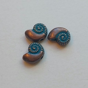 Op de afbeelding: Drie koperkleurige kralen met een spiraalvormig ontwerp en een turquoisekleurig midden. De kralen hebben de vorm van een nautilusschelp. De kralen zijn klein en hebben een gat om te rijgen.
