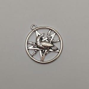 May include: A circular silver-toned metal pendant featuring a five-pointed star pentagram design. A detailed raven is positioned in the center of the star, flanked by two crescent moon shapes with decorative scrollwork accents.