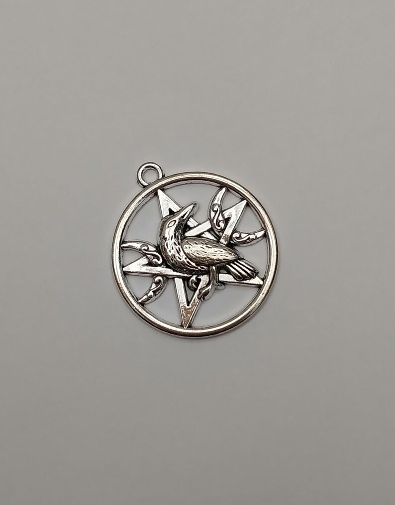 May include: A circular silver-toned metal pendant featuring a five-pointed star pentagram design. A detailed raven is positioned in the center of the star, flanked by two crescent moon shapes with decorative scrollwork accents.