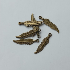 Pode incluir: Um conjunto de cinco pingentes de pena na cor bronze. Cada pingente apresenta um design detalhado de pena e uma pequena argola na parte superior para fixação. Estão espalhados sobre uma superfície branca.