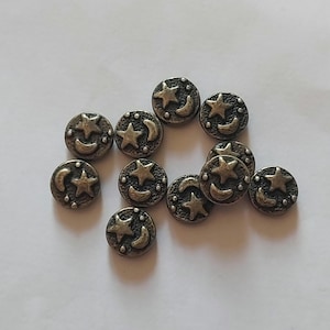 Könnte beinhalten: Eine Ansammlung von zehn runden Metallknöpfen, die jeweils ein erhabenes Reliefdesign eines fünfzackigen Sterns und einer Mondsichel aufweisen. Die Knöpfe haben ein dunkles, antiksilbernes Finish mit einer texturierten Oberfläche und kleinen Punkten.