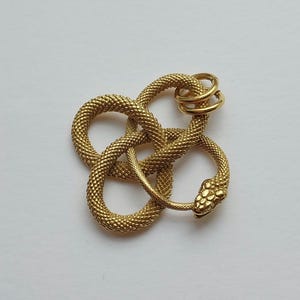 May include: A gold snake pendant with a textured, scale-like design. The snake is coiled in a figure-eight shape, with a detailed head and a small ring at the top. The jewellery piece is set against a white background.