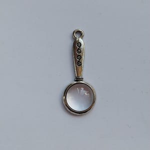Peut inclure: Charme de loupe en argent avec une lentille en verre transparent et une poignée décorative. La poignée présente un motif répétitif de cercles.