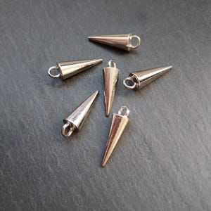 May include: Six silver-coloured cone-shaped metal studs with small loops at the top. The studs are arranged on a dark grey surface. These decorative studs are suitable for various craft and DIY projects.