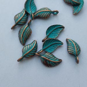 Peut inclure: Plusieurs breloques en forme de feuilles, de couleur turquoise et bronze, sont dispos&eacute;es sur une surface blanche. Les feuilles pr&eacute;sentent des d&eacute;tails de veines et une boucle pour l'enfilage. La couleur bronze est visible sur les bords et les nervures.