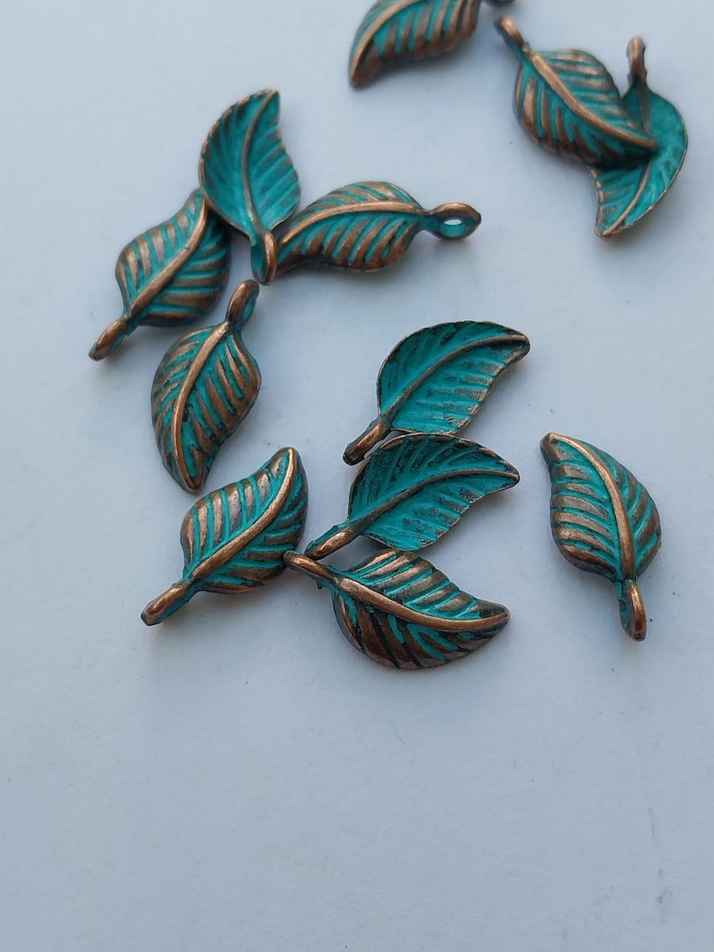 Peut inclure: Plusieurs breloques en forme de feuilles, de couleur turquoise et bronze, sont dispos&eacute;es sur une surface blanche. Les feuilles pr&eacute;sentent des d&eacute;tails de veines et une boucle pour l'enfilage. La couleur bronze est visible sur les bords et les nervures.