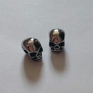 May include: Two small, polished silver-tone metal skull beads with detailed facial features including hollow eye sockets, a nasal cavity, and defined teeth. The surface of the beads is reflective, showing a smooth, metallic finish throughout.