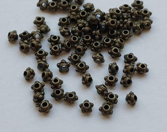 May include: A collection of small, bronze-colored metal beads. Each bead has a flower-like shape with six petals and a central hole. The beads are scattered on a white surface, suitable for jewelry making or crafts.