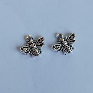 May include: Two silver-coloured bee charms. Each charm features detailed wings, body segments, and a small loop at the top for attaching to jewellery or other crafts. The bees are positioned against a plain white background.