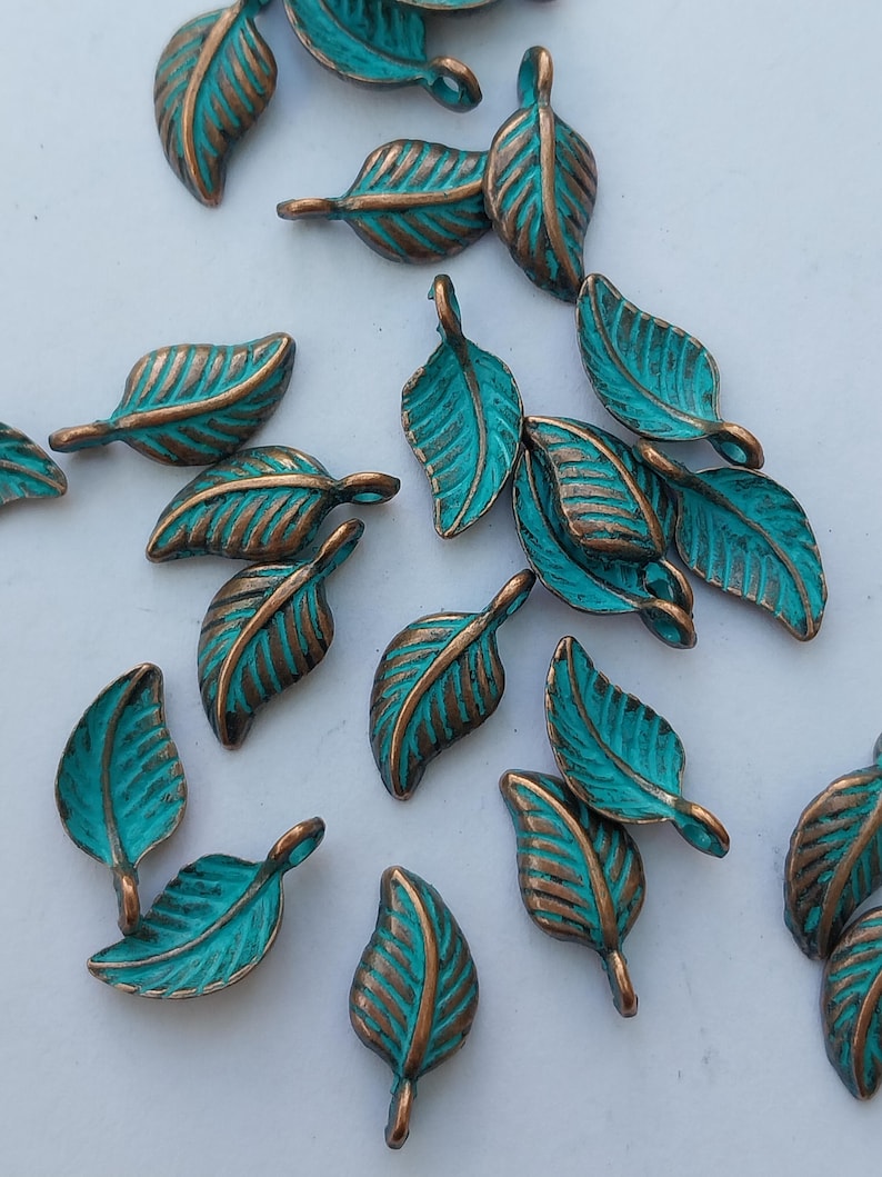 Peut inclure: Collection de breloques en forme de feuilles de couleur turquoise et bronze. Chaque feuille pr&eacute;sente un motif de veines d&eacute;taill&eacute; et une boucle pour l'attacher &agrave; des bijoux ou autres cr&eacute;ations. Les feuilles sont &eacute;parpill&eacute;es sur une surface blanche.