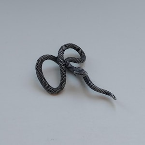 Pode incluir: Uma estatueta de cobra em metal escuro com uma superfície texturizada semelhante a escamas. A cobra está enrolada num laço com a cabeça a descansar perto do centro do corpo, apresentando olhos detalhados e uma cauda pontiaguda.