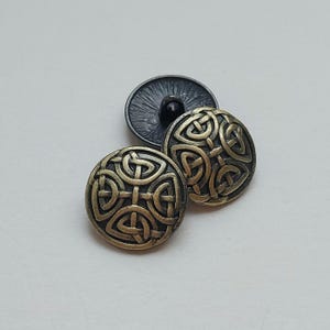 May include: Three antique brass-colored buttons with a Celtic knot design. Two buttons are face up, displaying the intricate knotwork, while the third is turned over, revealing a dark gray shank. The buttons are round and have a slightly aged appearance.