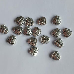 May include: A collection of small, silver-colored metal beads shaped like oak leaves. Each bead features a raised, textured vein pattern on the surface, with a metallic finish that reflects light. These individual leaf-shaped charms are scattered.