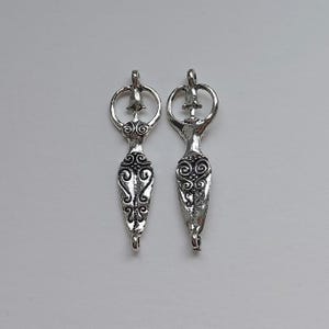 Peut inclure: Deux pendentifs argentés en forme de femme stylisée. Chaque pendentif présente une boucle en haut, une tête circulaire et un corps orné de volutes. Les pendentifs sont symétriques et d'aspect vintage.