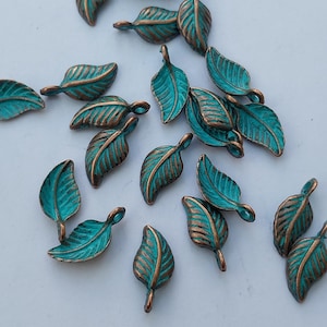 Peut inclure: Collection de petits charmes en m&eacute;tal en forme de feuilles. Chaque charme pr&eacute;sente une surface de couleur sarcelle avec des veines et des bords de couleur bronze. Les charmes sont dispers&eacute;s sur une surface blanche, cr&eacute;ant une esth&eacute;tique naturelle et organique.