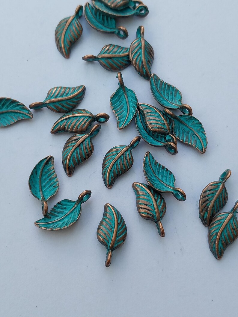 Peut inclure: Collection de petits charmes en m&eacute;tal en forme de feuilles. Chaque charme pr&eacute;sente une surface de couleur sarcelle avec des veines et des bords de couleur bronze. Les charmes sont dispers&eacute;s sur une surface blanche, cr&eacute;ant une esth&eacute;tique naturelle et organique.