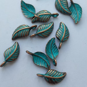 Peut inclure: Un ensemble de breloques en forme de feuilles en m&eacute;tal bronze avec une patine turquoise. Les feuilles pr&eacute;sentent des nervures d&eacute;taill&eacute;es et sont dispers&eacute;es sur une surface blanche. Id&eacute;al pour la cr&eacute;ation de bijoux ou les projets d'artisanat.
