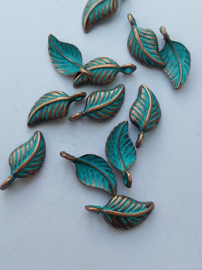 Peut inclure: Un ensemble de breloques en forme de feuilles en m&eacute;tal bronze avec une patine turquoise. Les feuilles pr&eacute;sentent des nervures d&eacute;taill&eacute;es et sont dispers&eacute;es sur une surface blanche. Id&eacute;al pour la cr&eacute;ation de bijoux ou les projets d'artisanat.