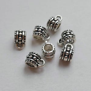 May include: Close-up of several small, silver-colored metal beads. Each bead is shaped like a miniature mug with a handle and is decorated with a series of small, raised dots and horizontal bands. The beads are scattered on a light-colored surface.