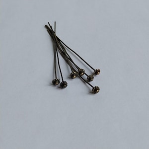 Könnte beinhalten: Ein Set aus sechs dünnen Metall-Kopfstiften mit antikem Bronze-Finish. Jeder Stift verfügt über eine kleine, dekorative, texturierte blumenförmige Kappe am oberen Ende, während die langen, geraden Schäfte schlicht und schlank gehalten sind.