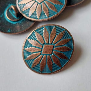 May include: Close-up of several copper-coloured buttons with a turquoise blue background. Each button features a central square design with radiating sunburst patterns. The buttons have a vintage, weathered appearance, suggesting they are antique or vintage.
