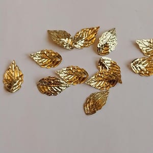 May include: A collection of small, gold-coloured metal leaf charms. Each charm features a detailed, embossed vein pattern on the surface and a small hole at the top for attachment. The leaves are arranged in a scattered pattern, showing various orientations and angles of the metallic, reflective finish.