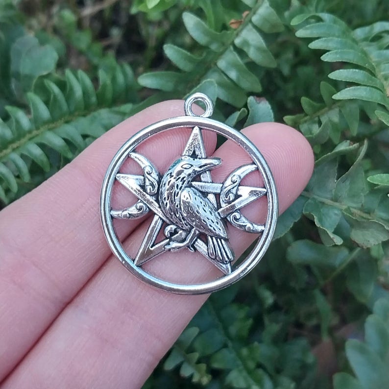 May include: A circular silver-toned metal pendant featuring a central five-pointed star, a detailed raven perched on the star, and two crescent moon shapes integrated into the design, with a small loop at the top for attachment.